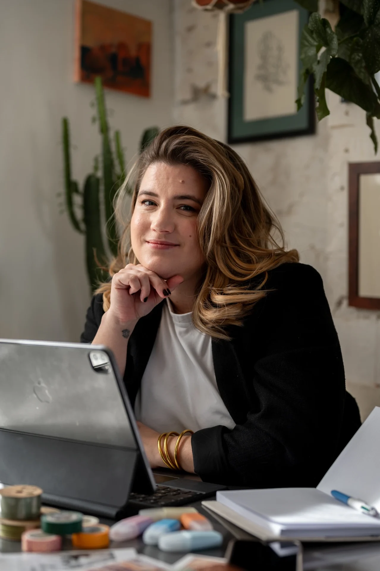 Élise en veste noire, le menton posé sur la main, assise à un bureau avec une tablette et un carnet ouvert.