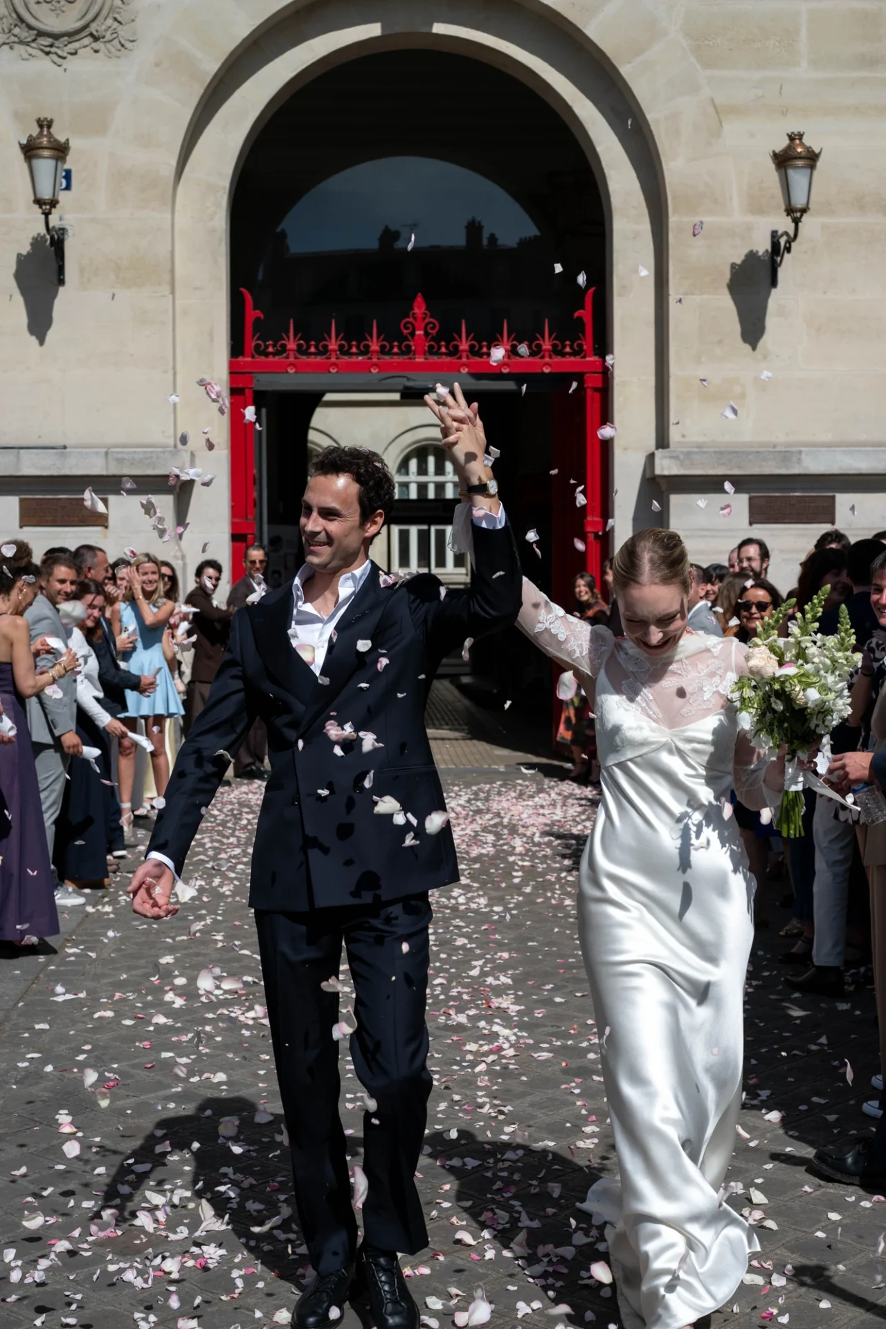 Full-length view of Louise and Lucas walking through falling petals, with applauding guests outside the town hall.